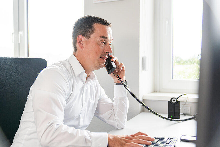 Konzentrierter Büroangestellter am Telefon mit Blick auf den Bildschirm und einer Tastatur vor sich.