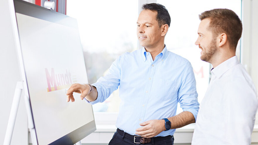 Zwei Männer stehen vor einer Tafel auf dem das Firmenlogo drauf ist