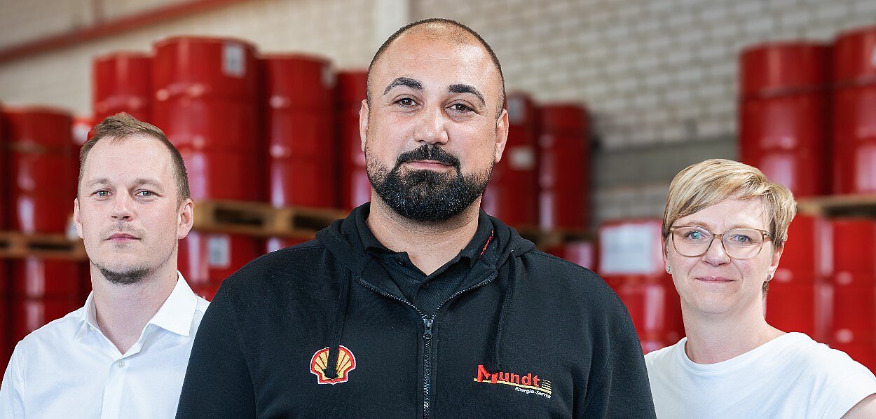 Zwei Männer und eine Frau stehen vor vielen Ölfässern