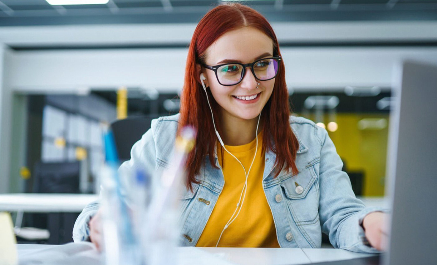 Eine lächelnde Frau die mit Kopfhörern im Ohr vor einem Computer sitzt